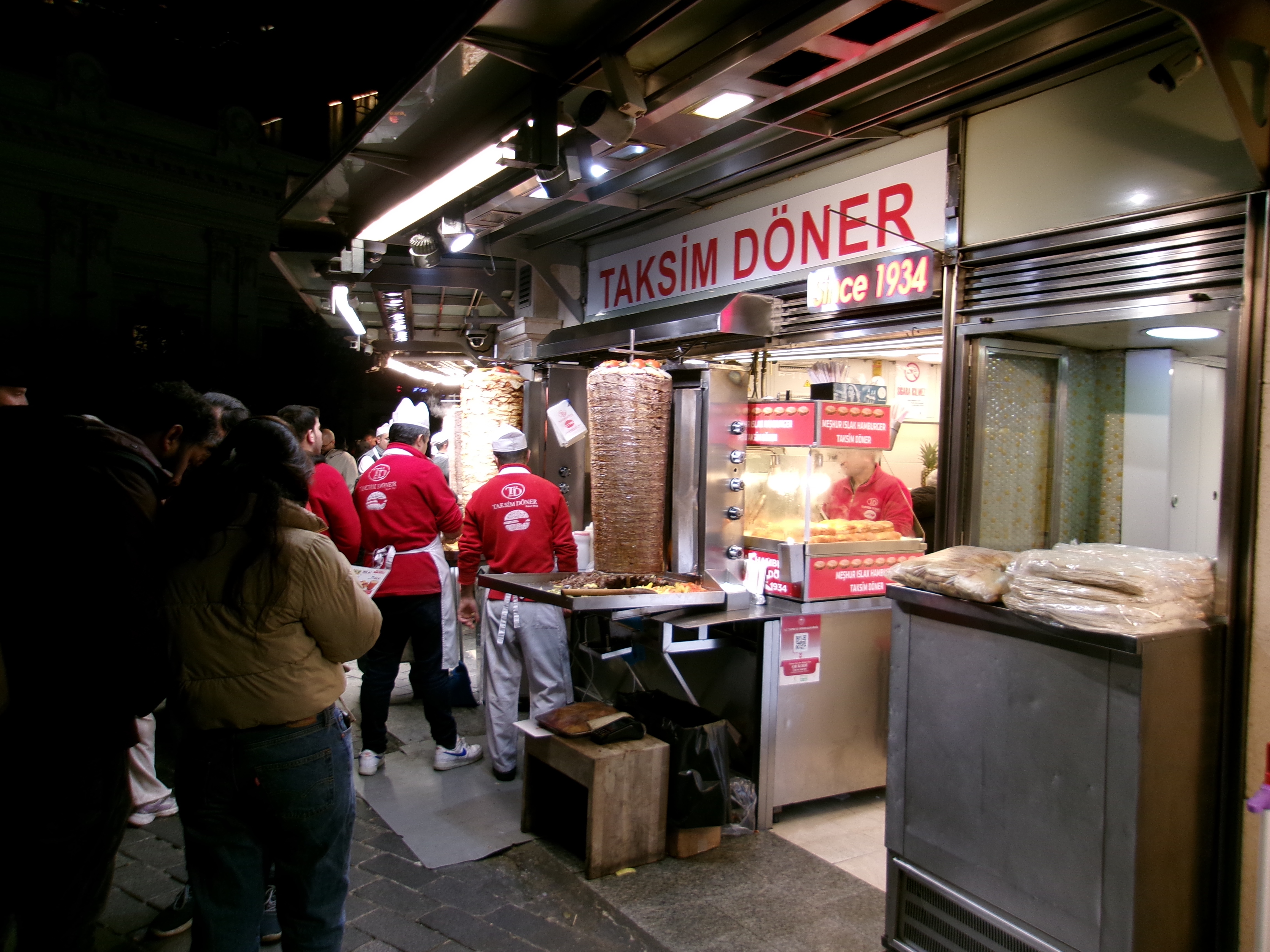 Busy Taksim Döner stall at night with vertical rotisseries and customers on cobblestones